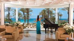 Elegante Hotellounge mit Flügel, gemütlichen Sesseln und Panoramablick auf Palmen, Terrasse und das türkisblaue Meer.