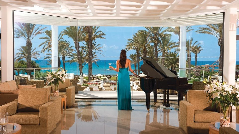 Elegante Hotellounge mit Flügel, gemütlichen Sesseln und Panoramablick auf Palmen, Terrasse und das türkisblaue Meer.