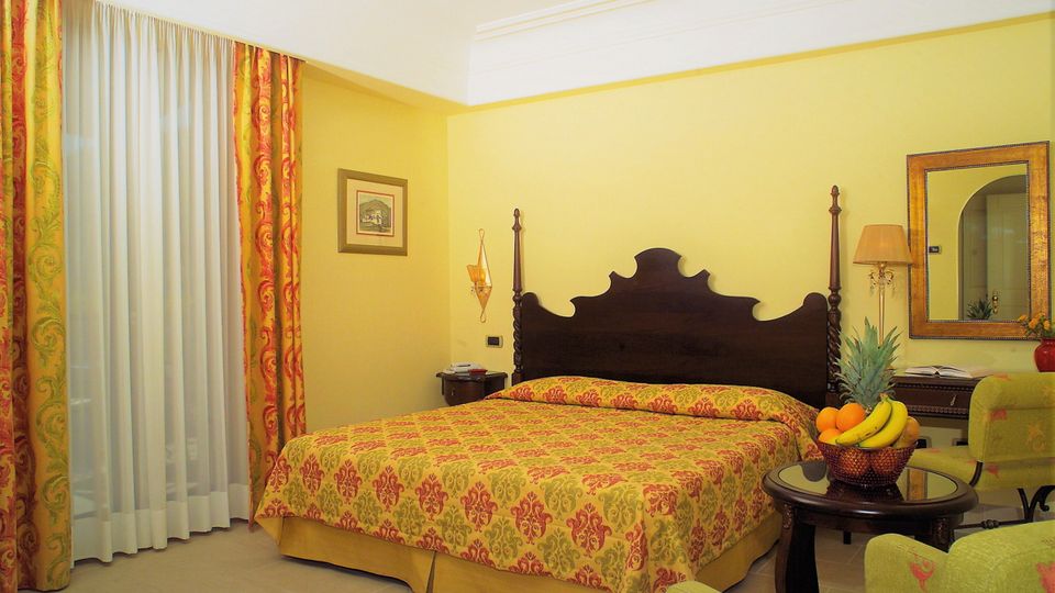 Klassisch eingerichtetes Hotelzimmer mit großem Doppelbett, floralen Vorhängen und stilvollen Möbeln in warmen Gelb- und Rottönen – auf dem Tisch eine Obstschale mit frischen Früchten.
