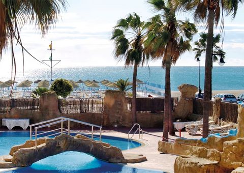 Poollandschaft mit Felsen, Palmen und kleiner Brücke, im Hintergrund Sandstrand mit Sonnenschirmen und Blick auf das glitzernde Meer unter blauem Himmel.