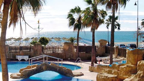 Poollandschaft mit Felsen, Palmen und kleiner Brücke, im Hintergrund Sandstrand mit Sonnenschirmen und Blick auf das glitzernde Meer unter blauem Himmel.