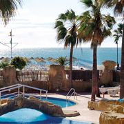 Poollandschaft mit Felsen, Palmen und kleiner Brücke, im Hintergrund Sandstrand mit Sonnenschirmen und Blick auf das glitzernde Meer unter blauem Himmel.