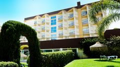 Gepflegte Gartenanlage mit geschnittenen Hecken, Palmen und einem grünen Torbogen vor einem mehrstöckigen Hotelgebäude mit Balkonen, im Hintergrund strahlend blauer Himmel.
