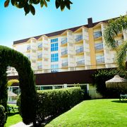 Gepflegte Gartenanlage mit geschnittenen Hecken, Palmen und einem grünen Torbogen vor einem mehrstöckigen Hotelgebäude mit Balkonen, im Hintergrund strahlend blauer Himmel.