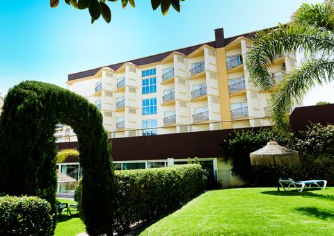 Gepflegte Gartenanlage mit geschnittenen Hecken, Palmen und einem grünen Torbogen vor einem mehrstöckigen Hotelgebäude mit Balkonen, im Hintergrund strahlend blauer Himmel.
