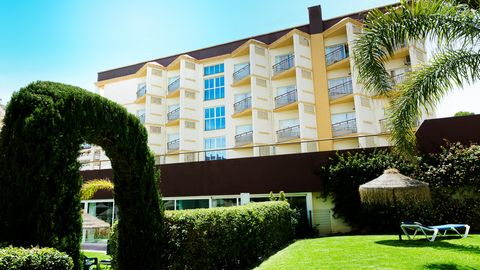 Gepflegte Gartenanlage mit geschnittenen Hecken, Palmen und einem grünen Torbogen vor einem mehrstöckigen Hotelgebäude mit Balkonen, im Hintergrund strahlend blauer Himmel.