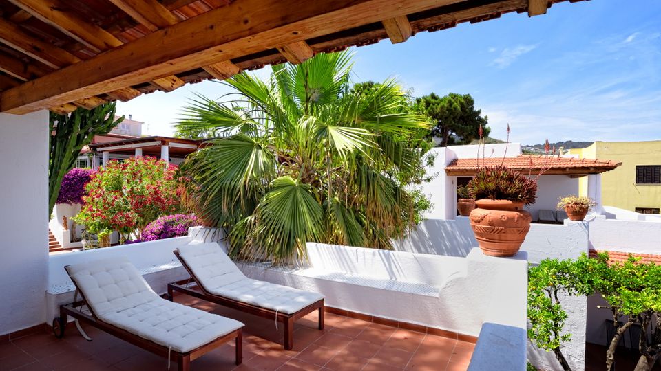 Überdachte Terrasse mit zwei gepolsterten Sonnenliegen, umgeben von Palmen, blühenden Pflanzen und weiß verputzten Mauern – mediterrane Ruheoase mit traditionellem Flair.