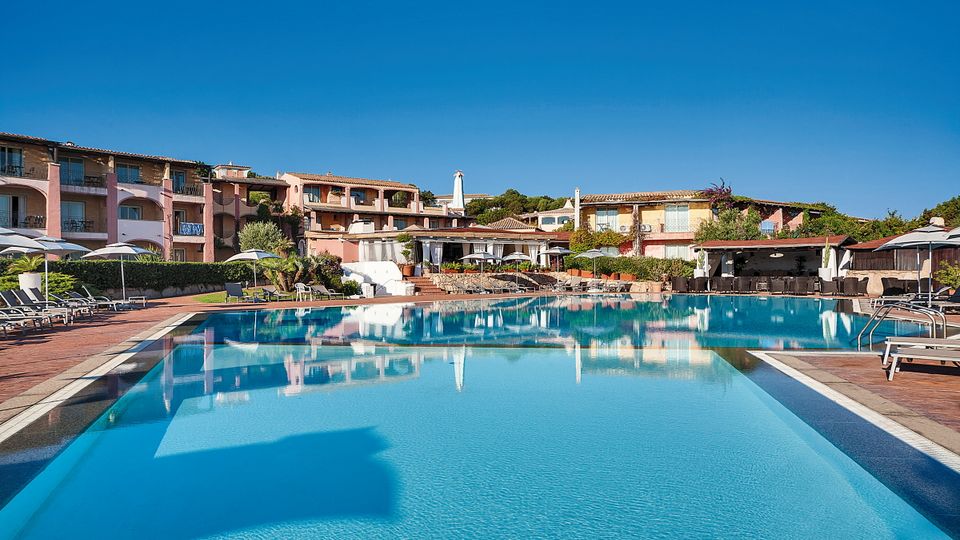 Weitläufige Poollandschaft eines mediterranen Resorts mit Sonnenliegen und Sonnenschirmen – dahinter bunte, terrakottafarbene Hotelgebäude mit Balkonen und üppiger Vegetation unter strahlend blauem Himmel.