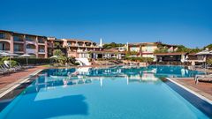Weitläufige Poollandschaft eines mediterranen Resorts mit Sonnenliegen und Sonnenschirmen – dahinter bunte, terrakottafarbene Hotelgebäude mit Balkonen und üppiger Vegetation unter strahlend blauem Himmel.