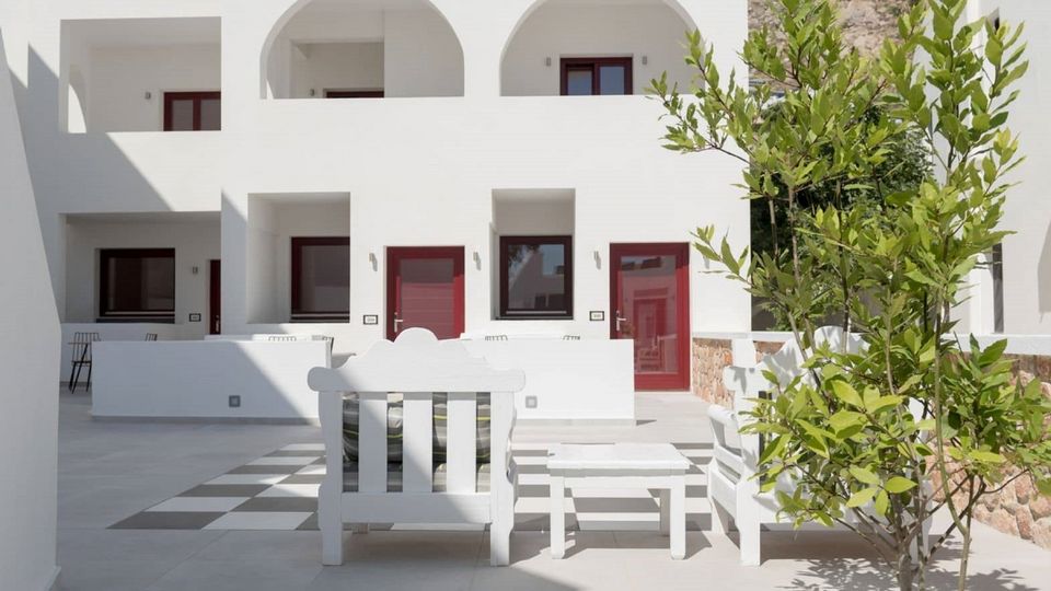 Lichter Innenhof mit weißen Sitzgelegenheiten im traditionellen Stil, rot umrahmte Türen und mediterraner Pflanzenakzent – perfekte Kulisse zum Verweilen und Entspannen.