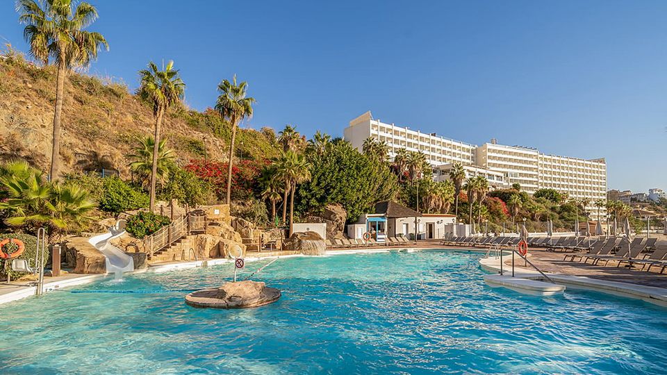 Weitläufige Poolanlage mit Wasserrutsche, Felsenlandschaft und Liegestühlen, umgeben von tropischen Palmen und blühender Vegetation, im Hintergrund ein großes Hotelgebäude an einem sonnigen Hang.