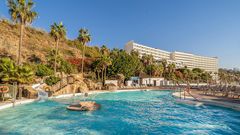 Weitläufige Poolanlage mit Wasserrutsche, Felsenlandschaft und Liegestühlen, umgeben von tropischen Palmen und blühender Vegetation, im Hintergrund ein großes Hotelgebäude an einem sonnigen Hang.