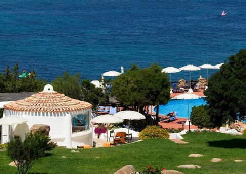 Blick auf einen idyllischen Hotelgarten mit mediterranen Pflanzen, Poolbereich mit Sonnenliegen und weißen Sonnenschirmen – im Hintergrund das tiefblaue Meer, das zum Träumen einlädt.