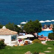 Blick auf einen idyllischen Hotelgarten mit mediterranen Pflanzen, Poolbereich mit Sonnenliegen und weißen Sonnenschirmen – im Hintergrund das tiefblaue Meer, das zum Träumen einlädt.