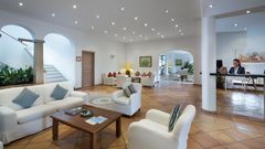 Helle Hotel-Lobby mit freundlicher Rezeption, gemütlichen Sitzgruppen in Weiß und mediterranem Fliesenboden, stilvoll dekoriert mit Pflanzen und Kunstwerken – ideal zum Entspannen beim Check-in.