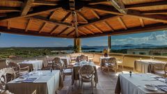 Elegantes Restaurant mit Holzdach und Panoramafenstern – gedeckte Tische mit Blick auf das Meer, grüne Landschaften und ferne Inseln unter blauem Himmel.