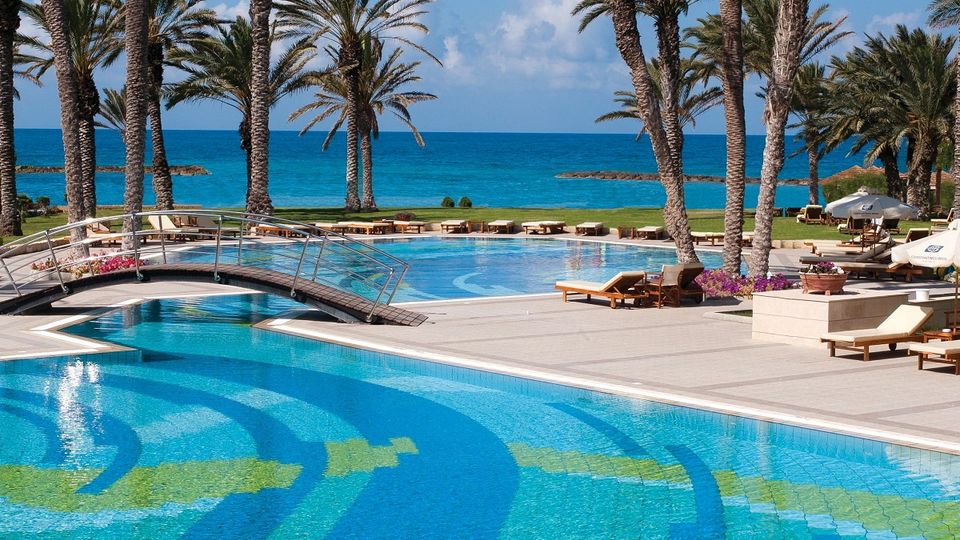 Ein geschwungenes Schwimmbecken mit türkisfarbenem Wasser und Mosaikmustern in verschiedenen Blau- und Grüntönen. Eine kleine Brücke führt über das Wasser, im Hintergrund reihen sich hohe Palmen vor dem azurblauen Meer.