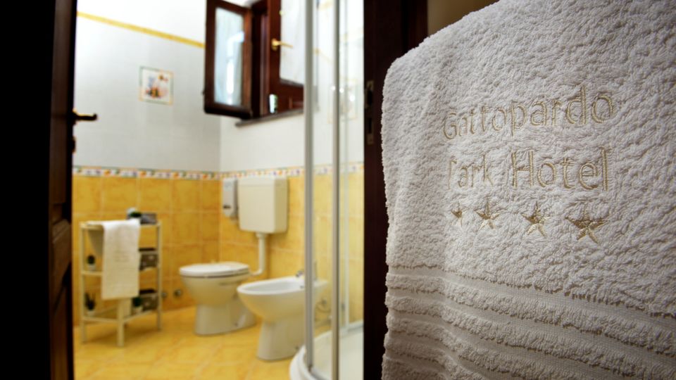 Helles Badezimmer mit gelb gefliesten Wänden und Dusche, im Vordergrund ein besticktes Handtuch mit dem Schriftzug „Gattopardo Park Hotel“ und vier Sternen.