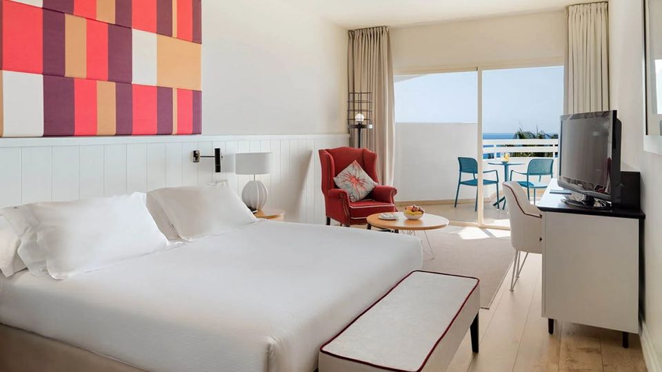 Modern eingerichtetes Hotelzimmer mit großem Doppelbett, stilvollem Sessel, Flachbild-TV und Zugang zu einem möblierten Balkon mit Meerblick.