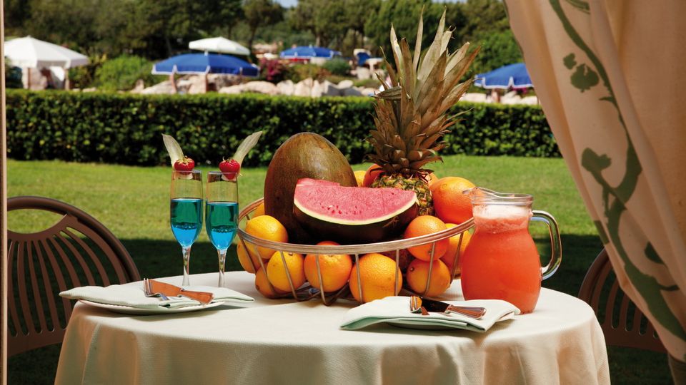 Stimmungsvoll gedeckter Tisch im Freien mit exotischer Obstschale, Wassermelone, Orangen und Ananas, daneben zwei blaue Cocktails und ein Krug frischer Saft – im Hintergrund Liegen mit blauen Sonnenschirmen auf einer grünen Wiese.