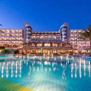 Beleuchtetes Hotel mit großem Poolbereich bei Nacht, eingerahmt von Palmen und klarer Abendstimmung.