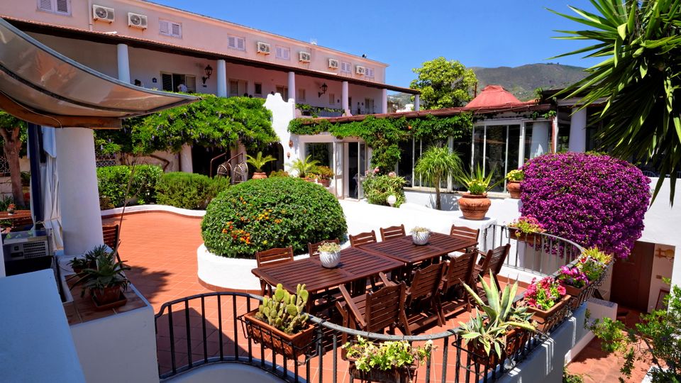 Charmanter Innenhof mit üppiger Bepflanzung, weiß verputzten Wänden und großer Terrasse mit Holzsitzgruppe – mediterranes Flair in ruhiger Umgebung mit Blick auf grüne Hügel.