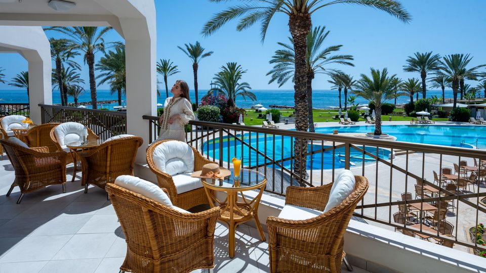 Terrasse mit gemütlichen Rattanmöbeln und Meerblick – im Hintergrund der Poolbereich, Palmen und das glitzernde Mittelmeer.