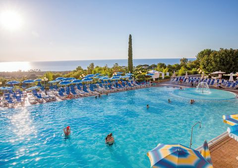Großer Außenpool mit Wasserspiel und vielen blauen Sonnenliegen und Schirmen – traumhafter Blick aufs Meer und ideale Bedingungen für entspannte Badetage unter der Sonne.