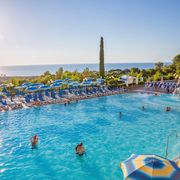 Großer Außenpool mit Wasserspiel und vielen blauen Sonnenliegen und Schirmen – traumhafter Blick aufs Meer und ideale Bedingungen für entspannte Badetage unter der Sonne.