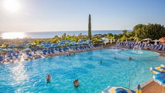 Großer Außenpool mit Wasserspiel und vielen blauen Sonnenliegen und Schirmen – traumhafter Blick aufs Meer und ideale Bedingungen für entspannte Badetage unter der Sonne.