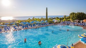 Großer Außenpool mit Wasserspiel und vielen blauen Sonnenliegen und Schirmen – traumhafter Blick aufs Meer und ideale Bedingungen für entspannte Badetage unter der Sonne.