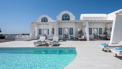 Modernes Boutique-Hotel im kykladischen Stil mit weißer Fassade, blauen Fensterläden und Poolterrasse mit Liegen – klare Architektur und mediterranes Flair unter strahlend blauem Himmel.