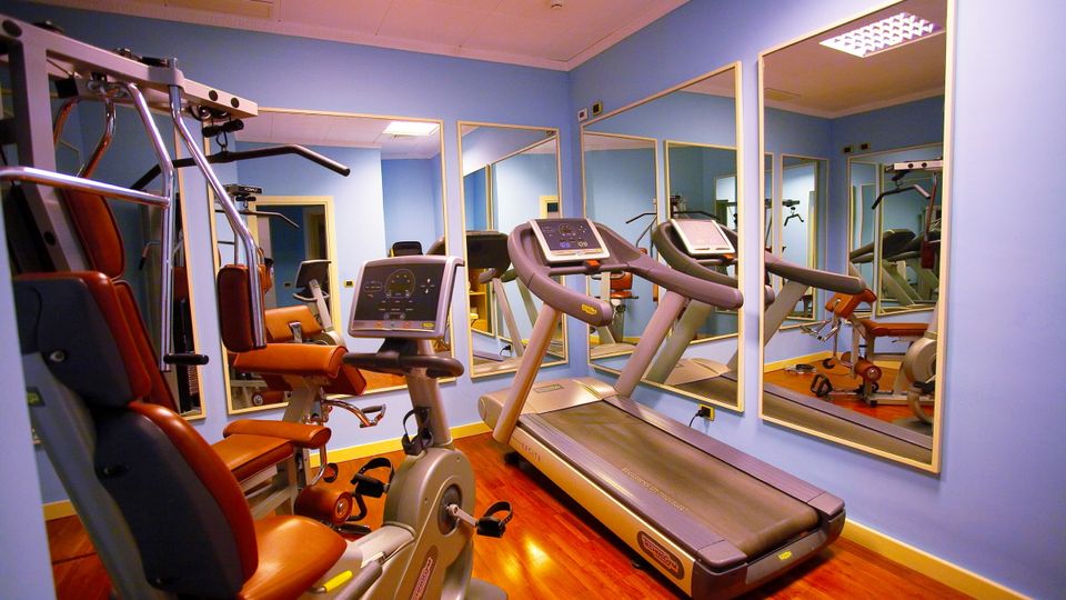 Kompakt ausgestatteter Fitnessraum mit Laufband, Ergometer, Kraftstation und großen Spiegeln – moderne Geräte auf glänzendem Holzfußboden in freundlicher Atmosphäre.