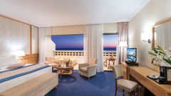 Helles, komfortables Doppelzimmer mit Sitzbereich, Balkon und traumhaftem Meerblick bei Sonnenuntergang.