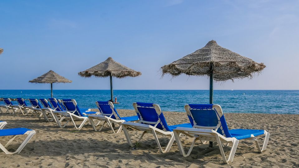 Reihe blauer Liegestühle mit Strohdach-Sonnenschirmen an einem Sandstrand, mit Blick auf das ruhige blaue Meer unter klarem Himmel.
