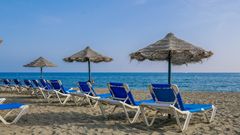 Reihe blauer Liegestühle mit Strohdach-Sonnenschirmen an einem Sandstrand, mit Blick auf das ruhige blaue Meer unter klarem Himmel.