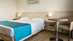 Helles Doppelzimmer mit Holzboden, schlichter Einrichtung und türkisfarbenem Bettläufer.